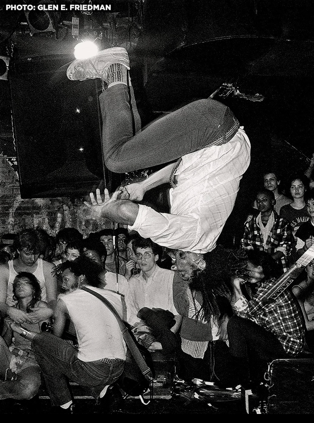 H.R. of Bad Brains mid-backflip on stage over a pressing crowd, black and white