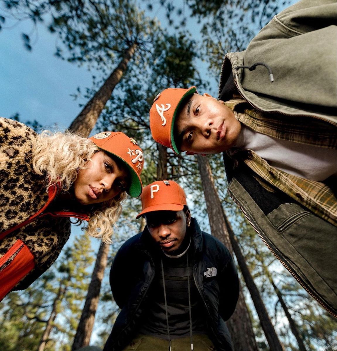 Three friends in orange caps among pines, shot from below